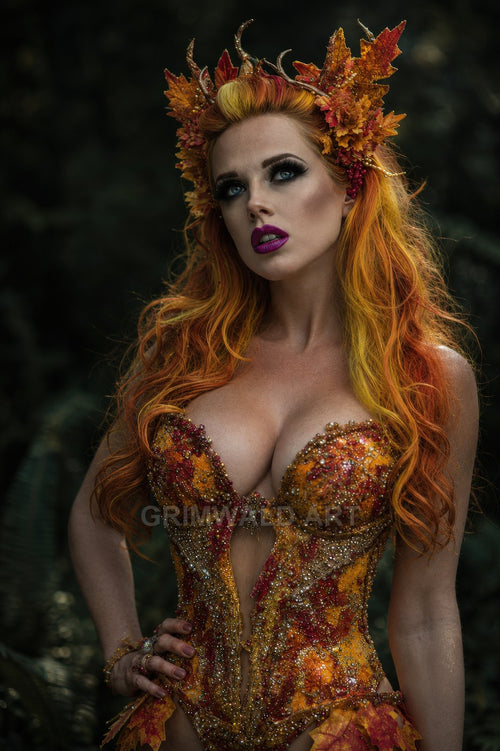 A fae woman with fiery orange hair wears an antlered leaf crown and jewel-encrusted autumn corset, posed in a dim forest setting.