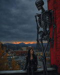 GRID THUMBNAIL — A gothic woman in black sits on a rooftop beside a towering skeleton, city lights below and fiery sunset beyond dark mountains.