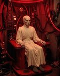 GRID THUMBNAIL — A bald robed figure sits in a red-lit industrial chair beneath a glowing neon ring, surrounded by cables, control panels, and machinery in a confined chamber.